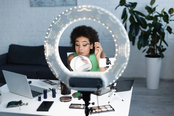 Jeune femme afro-américaine appliquant mascara près du miroir, des appareils et de la lumière anneau à la maison — Photo de stock