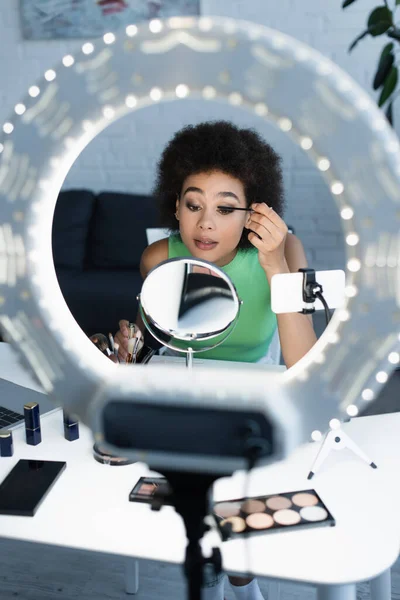 Blogger afroamericano che applica mascara vicino smartphone e ring light a casa — Foto stock