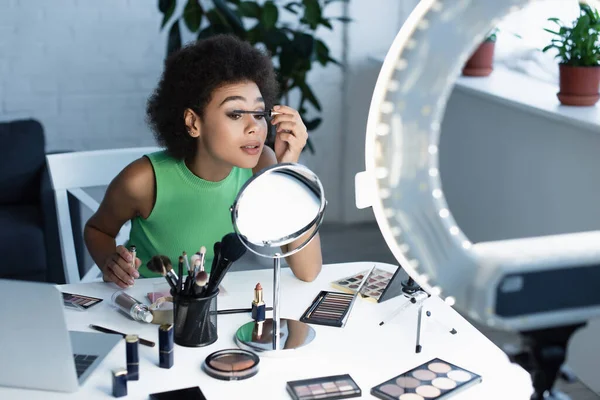 Jolie blogueuse afro-américaine appliquant du mascara près du miroir, des gadgets et des cosmétiques décoratifs à la maison — Photo de stock