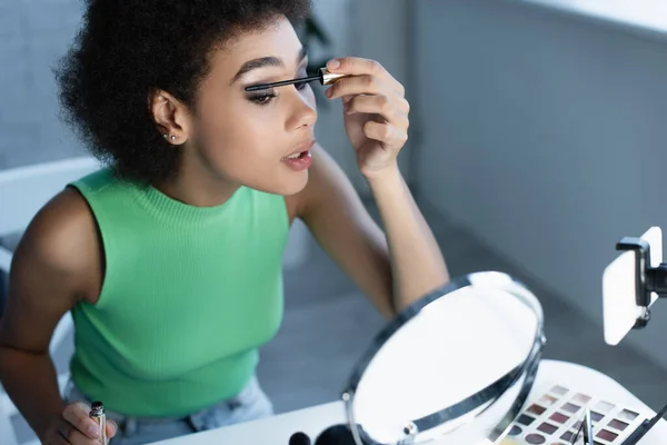 Blogueiro afro-americano aplicando rímel perto do espelho borrado e smartphone — Fotografia de Stock