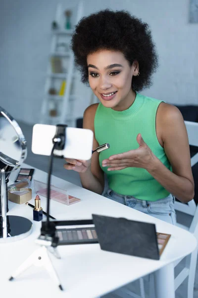 Blogueiro americano africano positivo segurando rímel perto de smartphone em casa — Fotografia de Stock