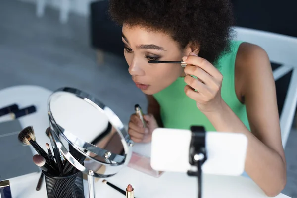 African american blogger applying mascara near cosmetic brushes and smartphone — Stock Photo