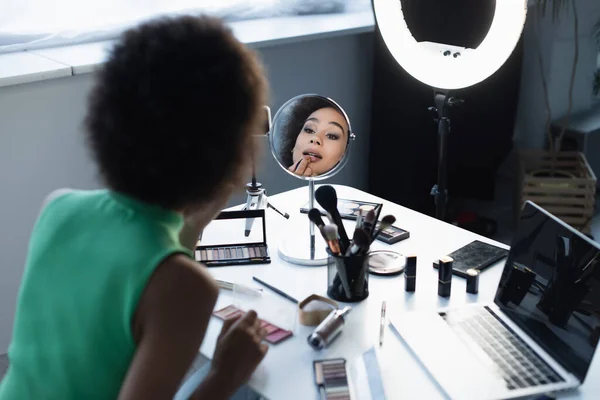 Blogueiro afro-americano desfocado aplicando cosméticos decorativos perto de gadgets e anel de luz em casa — Fotografia de Stock