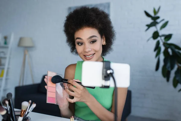 Blogger afroamericano sosteniendo rubor cerca de teléfono inteligente borroso en casa - foto de stock