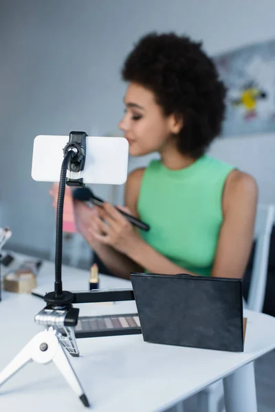 Smartphone près de cosmétiques décoratifs et femme afro-américaine floue à la maison — Photo de stock