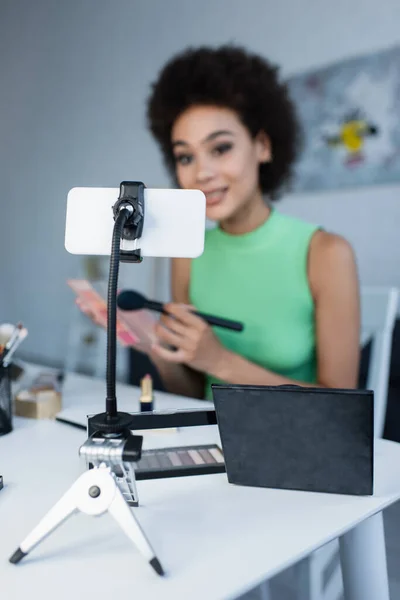 Teléfono móvil cerca de cosméticos decorativos y bloguero afroamericano borroso en casa - foto de stock