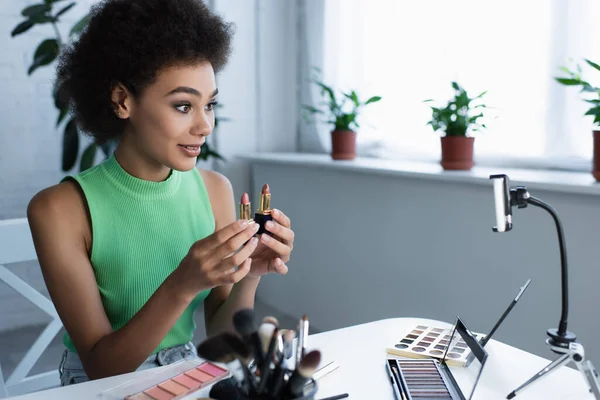 Sonriente blogger afroamericano sosteniendo lápices labiales cerca de cosméticos decorativos y teléfonos inteligentes - foto de stock