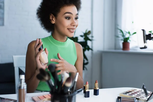 Mulher afro-americana positiva mostrando rosto fundação perto de cosméticos em casa — Fotografia de Stock