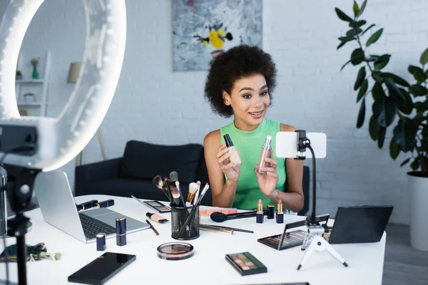 Smiling african american blogger holding face foundations near smartphone at home — Fotografia de Stock