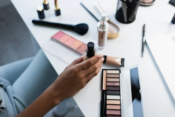 Vista recortada de la mujer afroamericana sosteniendo la base de la cara cerca de cosméticos borrosos en la mesa - foto de stock