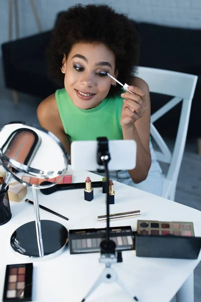 Mujer afroamericana feliz aplicando sombras de ojos cerca del espejo y el teléfono inteligente en casa - foto de stock