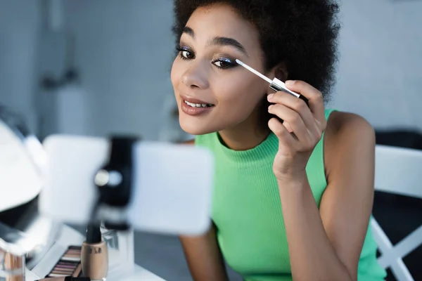 Blogger afroamericano positivo mirando el teléfono inteligente mientras aplica sombras de ojos - foto de stock
