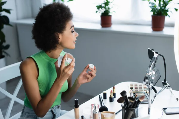 Vista laterale del blogger afro-americano che spruzza profumo vicino a cosmetici e smartphone a casa — Foto stock