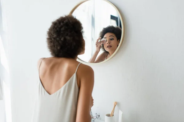 Young african american woman in silk nightgown wiping eye with cotton pad near mirror in bathroom — Fotografia de Stock