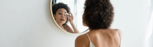 Femme afro-américaine essuyant les yeux avec coton près du miroir dans la salle de bain, bannière — Photo de stock