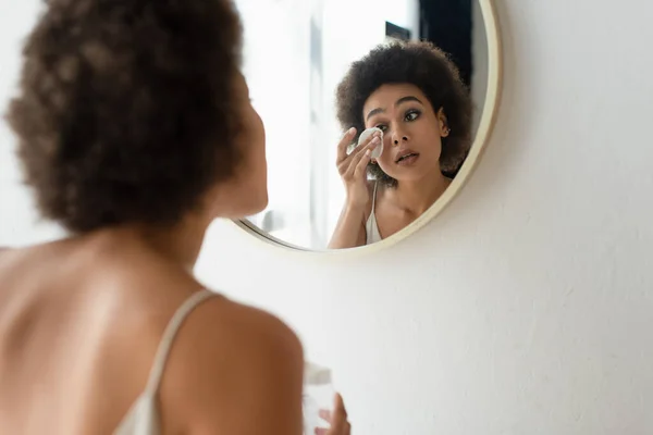 Mujer afroamericana borrosa limpiando la cara con almohadilla de algodón en el baño - foto de stock