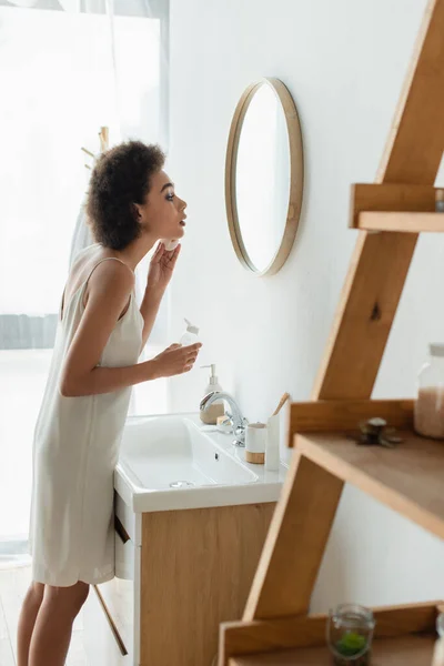 Vista laterale della giovane donna africana americana che pulisce il viso con un batuffolo di cotone in bagno — Foto stock