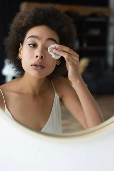 Jeune femme afro-américaine nettoyant les yeux avec du coton près du miroir dans la salle de bain — Photo de stock