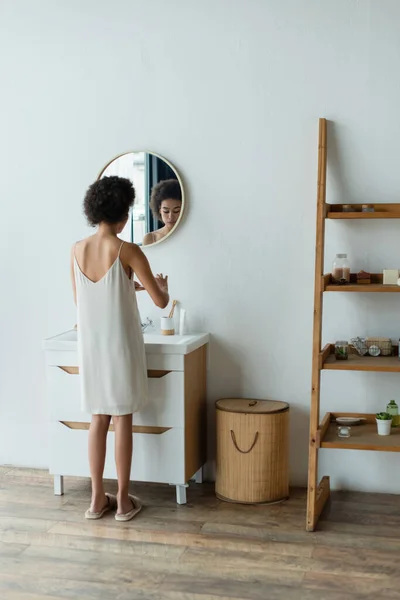 Mujer afroamericana en camisón de seda de pie cerca del lavabo y espejo en el baño - foto de stock