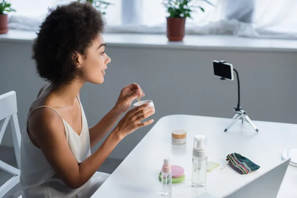 Side view of smiling african american blogger holding jar with eye patches near smartphone at home — Foto stock