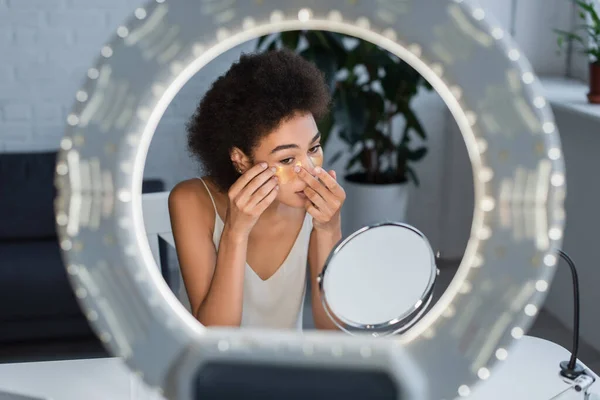 Mujer afroamericana joven aplicando parche para los ojos cerca del espejo y la luz del anillo en casa - foto de stock