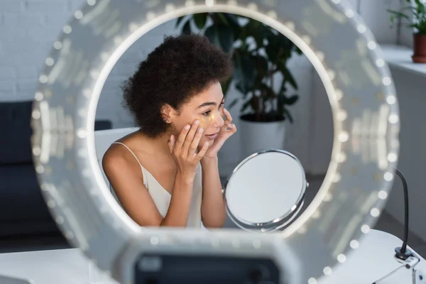 Mujer afroamericana que aplica parches en los ojos cerca del espejo y la luz borrosa del anillo en casa - foto de stock
