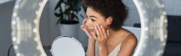 Mujer afroamericana joven que aplica parches para los ojos cerca del espejo y luz borrosa del anillo, pancarta - foto de stock