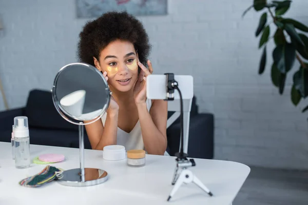 Blogueiro americano africano positivo aplicando manchas oculares perto do espelho e do smartphone em casa — Fotografia de Stock