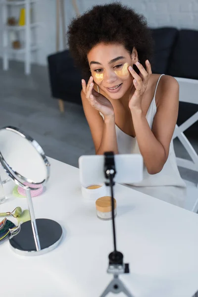 Cheerful african american blogger applying eye patches near blurred smartphone and mirror at home — Stockfoto