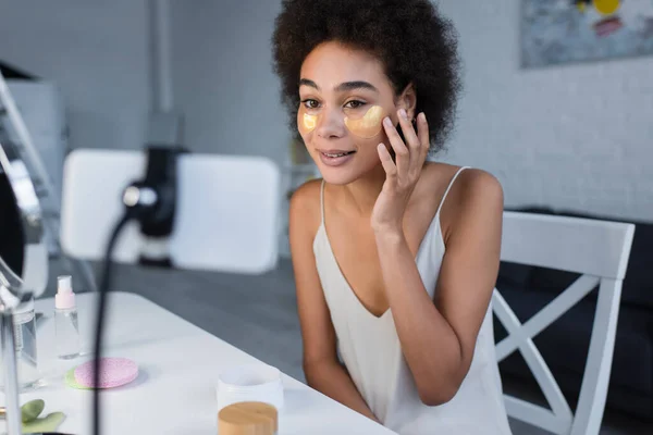 Young african american blogger applying eye patches near blurred cellphone at home — Fotografia de Stock