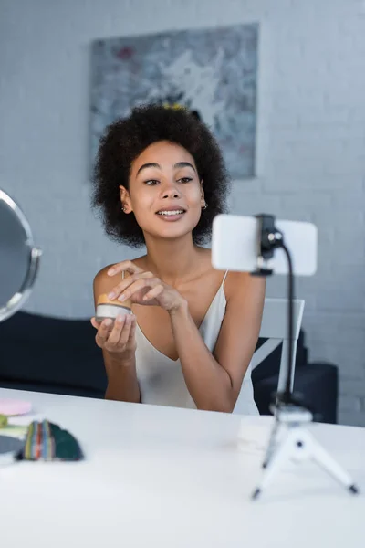 Sonriente blogger afroamericano sosteniendo crema cosmética cerca de teléfono inteligente y espejo en casa - foto de stock