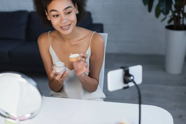 Alegre blogger afroamericano sosteniendo crema cosmética cerca del teléfono celular en casa - foto de stock