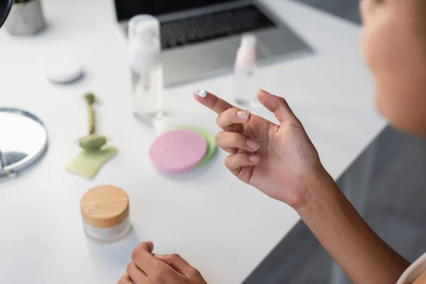 Vista ritagliata di donna afro-americana con crema cosmetica vicino al computer portatile offuscata e rullo di giada sul tavolo — Foto stock