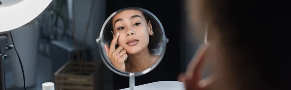 Mujer afroamericana joven aplicando crema cosmética cerca del espejo y la luz del anillo en casa, pancarta - foto de stock