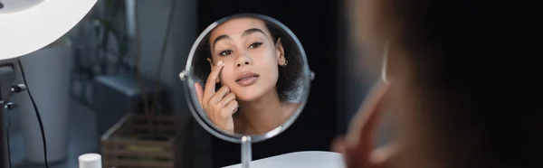 Jovem afro-americana aplicando creme cosmético perto do espelho e anel de luz em casa, banner — Fotografia de Stock