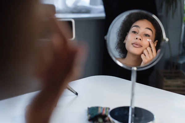 African american blogger applying cosmetic cream on face near mirror and smartphone at home — Fotografia de Stock