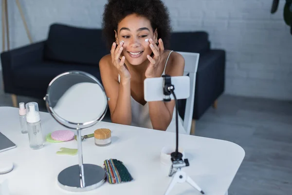 Cheerful african american blogger applying cosmetic cream near cellphone and jade roller at home — Fotografia de Stock