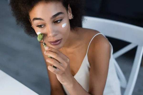 Femme afro-américaine avec crème cosmétique sur le visage à l'aide d'un rouleau de jade à la maison — Photo de stock