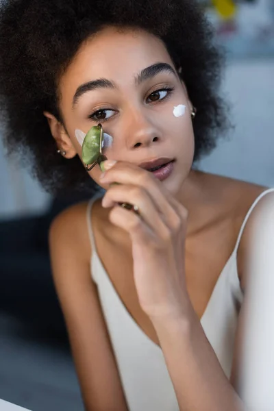 Femme afro-américaine appliquant crème cosmétique avec rouleau de jade à la maison — Photo de stock