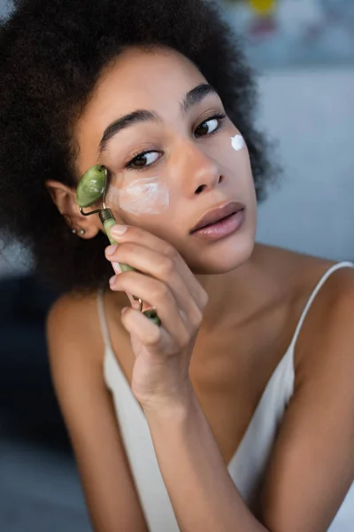 Jovem afro-americana aplicando creme cosmético com rolo de jade em casa — Fotografia de Stock