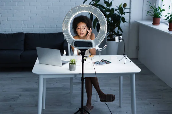 African american blogger applying cosmetic cream and looking at smartphone near laptop and ring light at home — Foto stock
