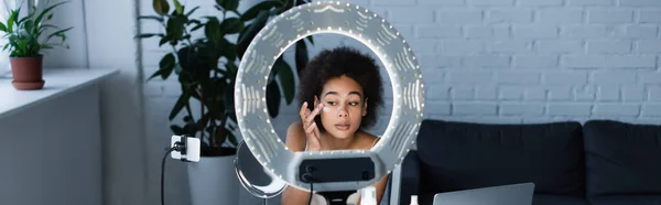 Mujer afroamericana aplicando crema cosmética en la cara cerca del teléfono celular y luz de anillo en casa, pancarta - foto de stock