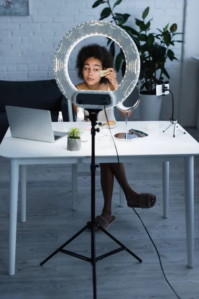 African american blogger applying cosmetic cream with gua sha near gadgets and ring light at home — Fotografia de Stock