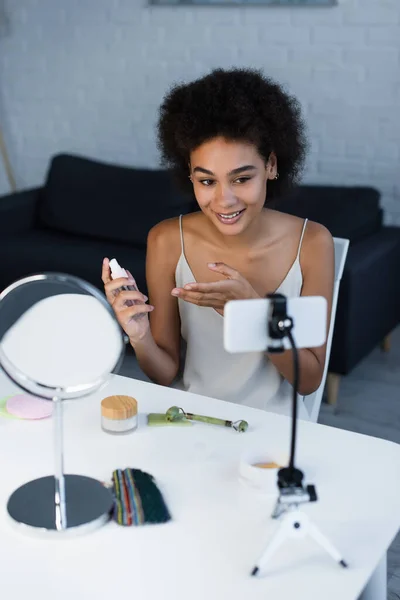 Sonriente blogger afroamericano apuntando a loción cosmética cerca de smartphone y espejo en casa - foto de stock