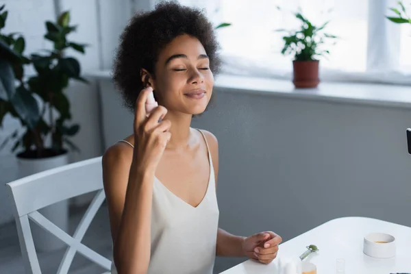 Joven blogger afroamericano aplicando loción en la cara cerca de parches para los ojos y rodillo de jade en casa - foto de stock