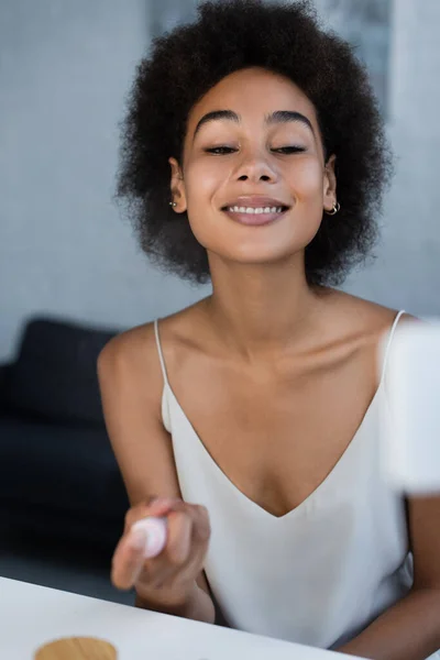 Sonriente mujer afroamericana sosteniendo loción cerca de un teléfono celular borroso en casa - foto de stock