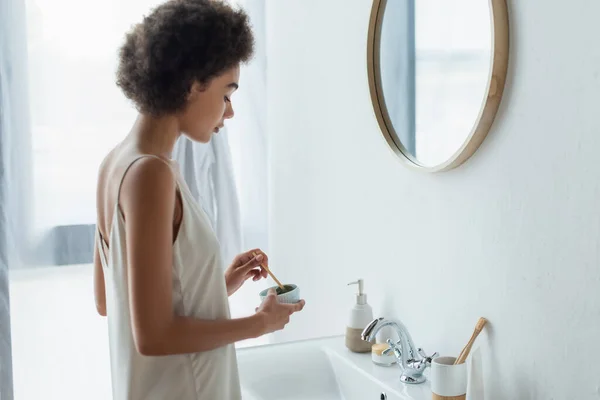 Vue latérale de la jeune femme afro-américaine mélangeant masque facial près du miroir dans la salle de bain — Photo de stock