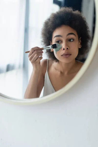 Femme afro-américaine appliquant masque facial avec pinceau cosmétique près du miroir à la maison — Photo de stock