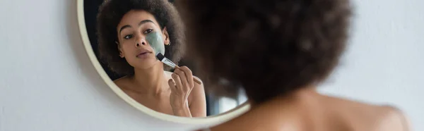 Junge afrikanisch-amerikanische Frau mit Kosmetikbürste, die Gesichtsmaske in der Nähe von Spiegel aufträgt, Banner — Stockfoto