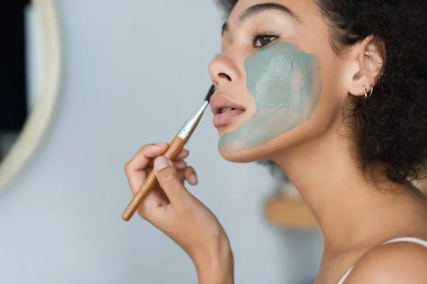 Jeune femme afro-américaine appliquant un masque d'argile sur le visage dans la salle de bain — Photo de stock